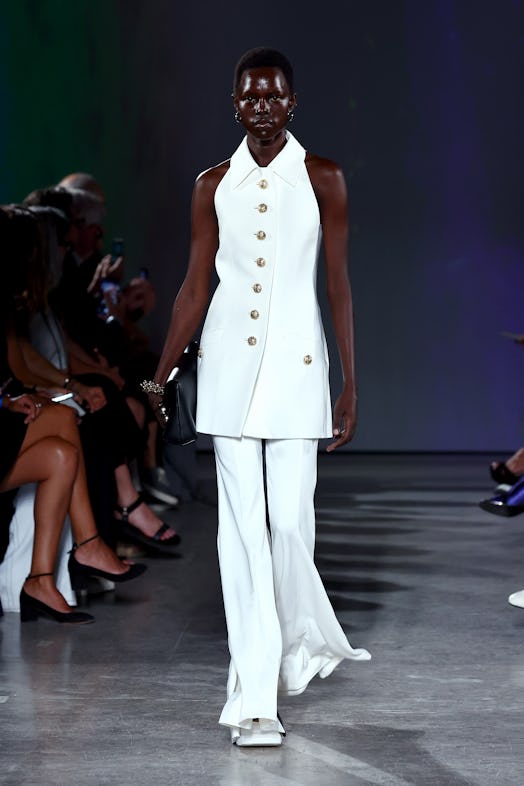 A model wears a white outfit Proenza Schouler, matching a gold-buttoned long waistcoat and wide, flo...