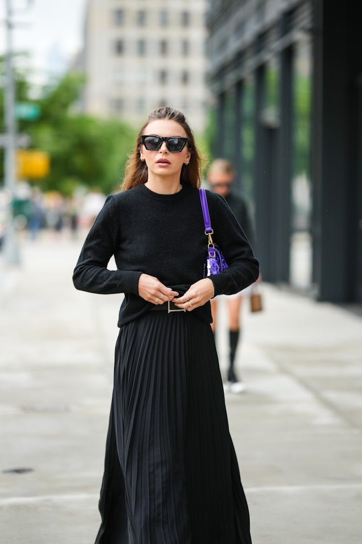 A guest wearing black sunglasses, a black pullover, a black pleated long skirt and a purple shiny se...