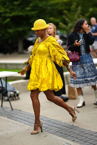 A guest wears a yellow mesh hat, earrings, a camel shiny leather crossbody bag, a gold shiny ruffled...