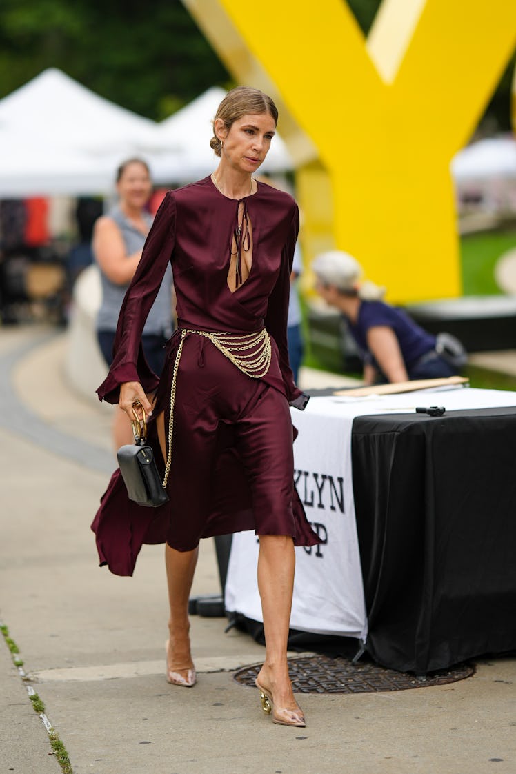 A guest wearing gold earrings, a chain pendant necklaces and a chain belt, a burgundy silk dress and...