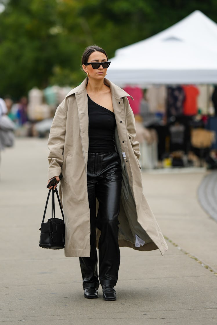 A guest wearing black sunglasses, a black asymmetrical shoulder top, a beige trench coat and black s...