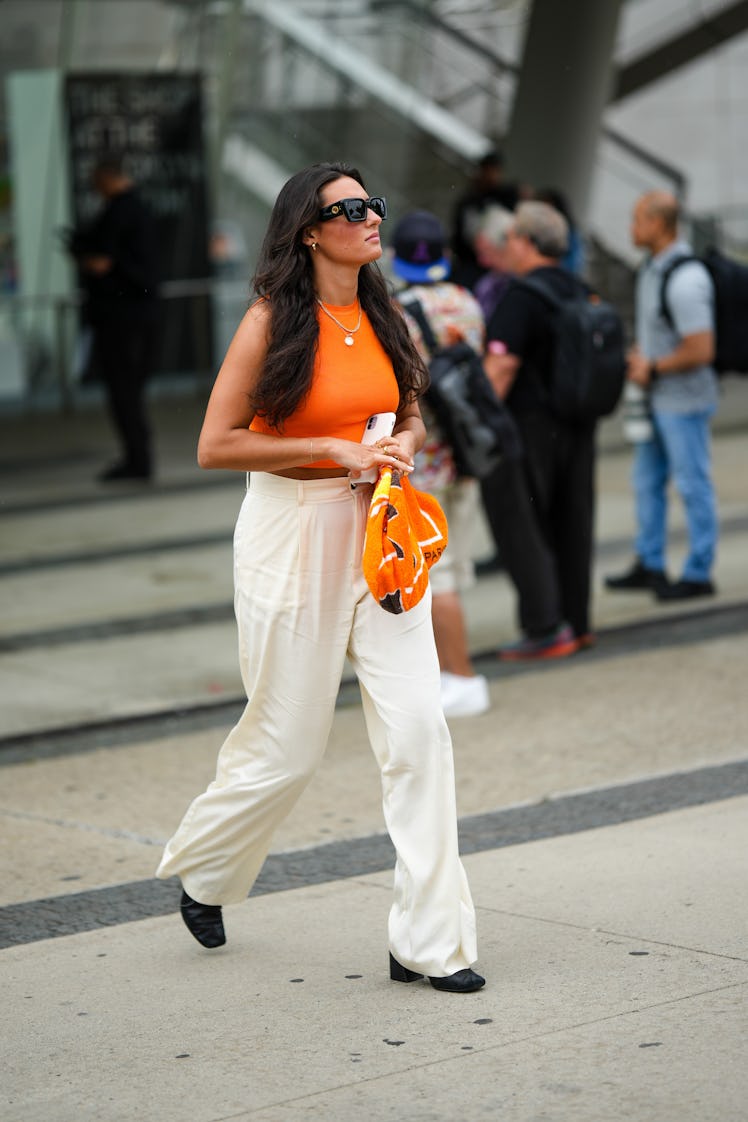 A guest wears black sunglasses, a neon orange halter neck cropped top, a gold chain necklace, gold e...