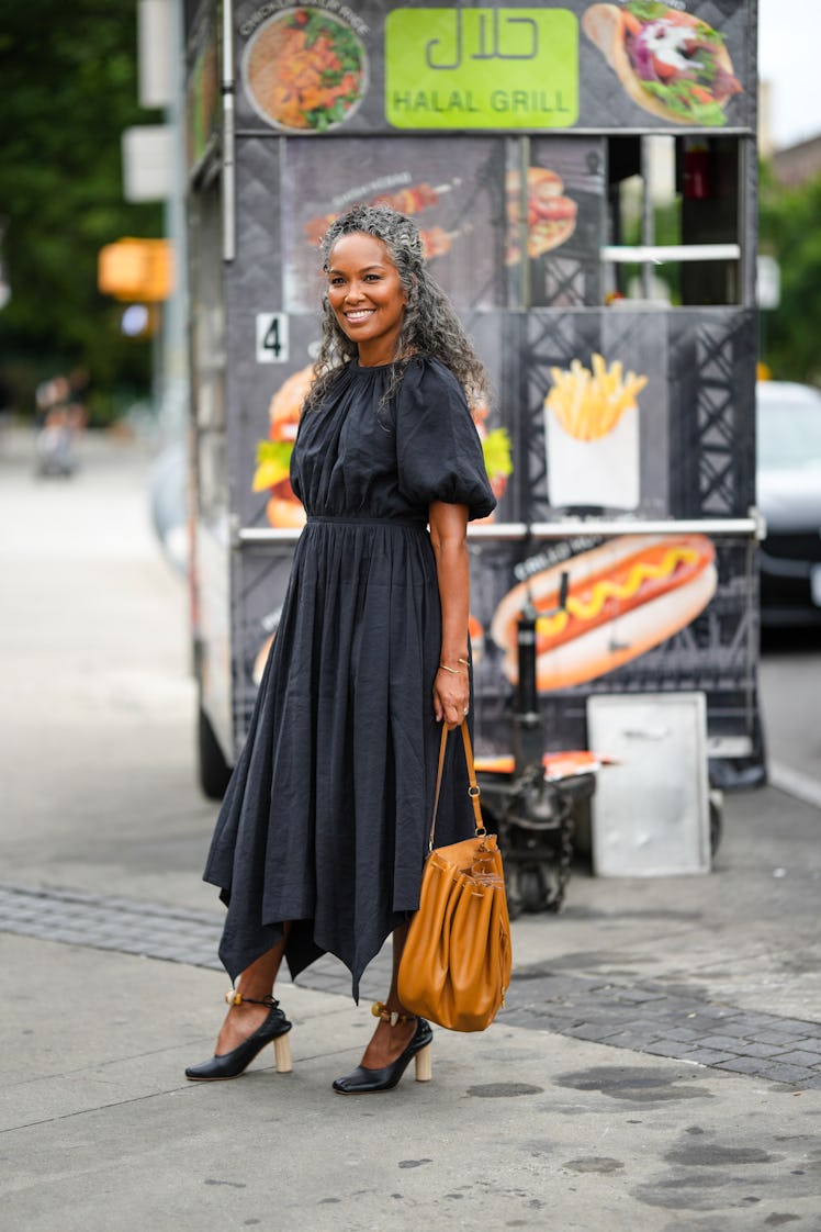 A guest wearing a dark gray pleated long dress and a camel shiny pleated bag with black heels shoes.