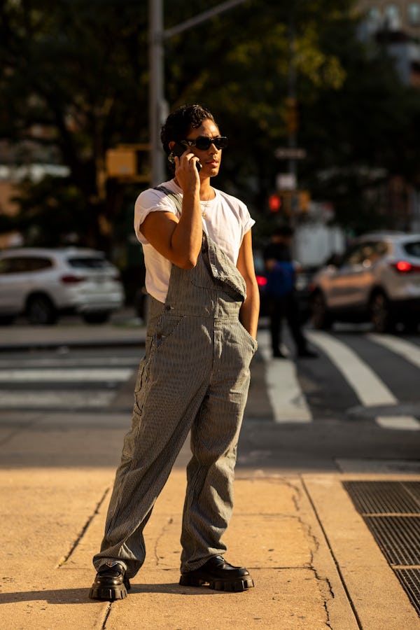 A man in a white shirt and grey overalls speaking on his phone