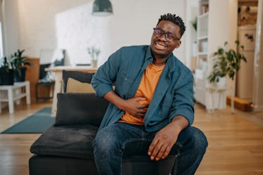 A man sits on a couch, clutching his stomach in pain.