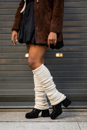 A woman in a black mini dress, brown jacket, white bag, black shoes and white leg warmers close-up