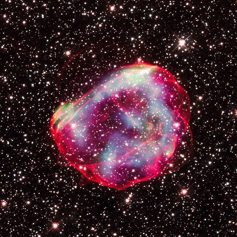 Glowing pink, green, and blue dust clouds make up a supernova remnant against a backdrop of bright s...