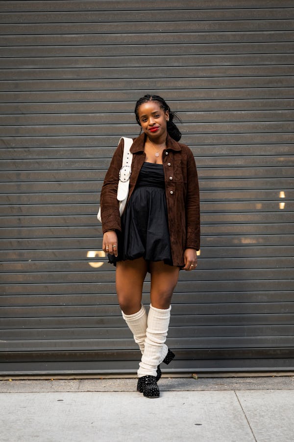 A woman in a black mini dress, brown jacket, white bag, black shoes and white leg warmers