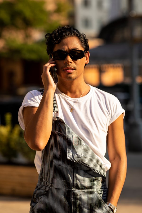 A man in a white shirt and grey overalls speaking on his phone close-up