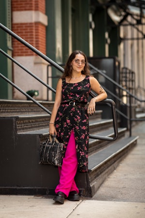 A woman wearing a floral dress over pink pants, a black bag and black platform shoes standing