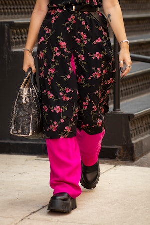 A woman wearing a floral dress over pink pants, a black bag and black platform shoes walking