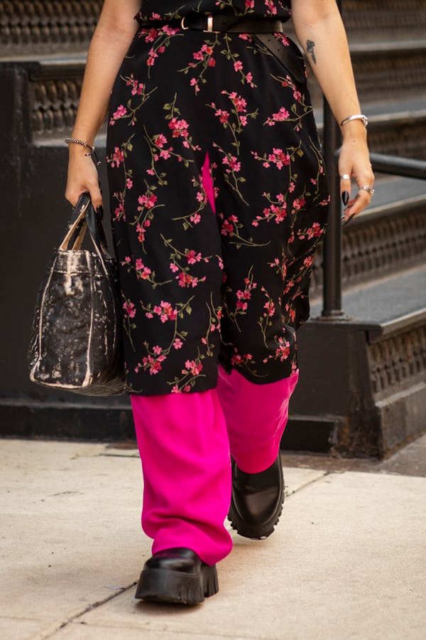 A woman wearing a floral dress over pink pants, a black bag and black platform shoes walking