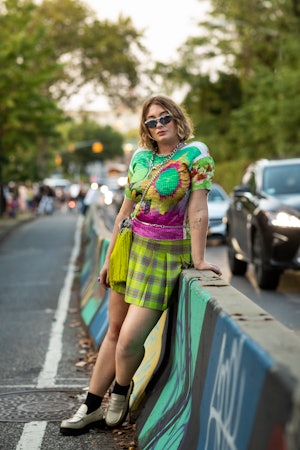 A woman in a floral multi-colored top, green plaid skirt, beige shoes and dad sunglasses