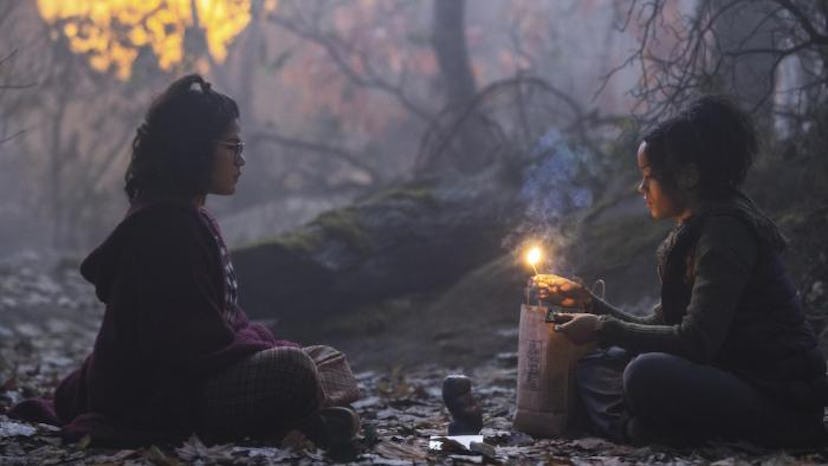 Two girls sit in the woods with a candle between them.
