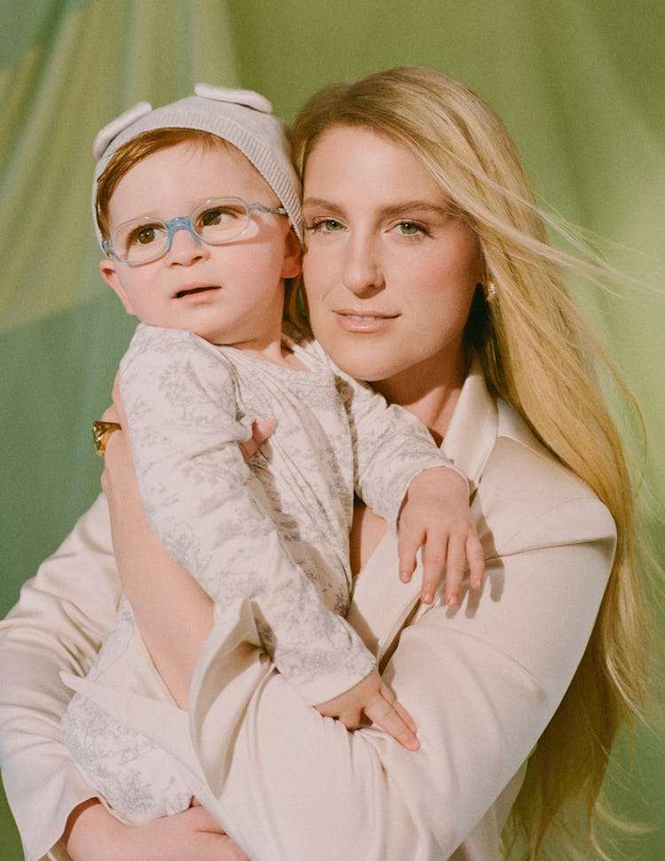 Meghan Trainor stands in front of green curtains, looking forward while holding her son, both dresse...
