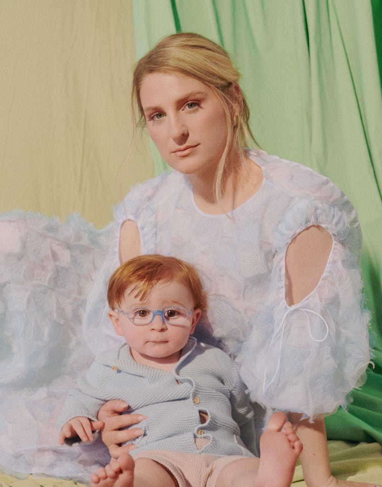 Meghan Trainor sitting on the ground in a white dress, holding her son while they both look straight...