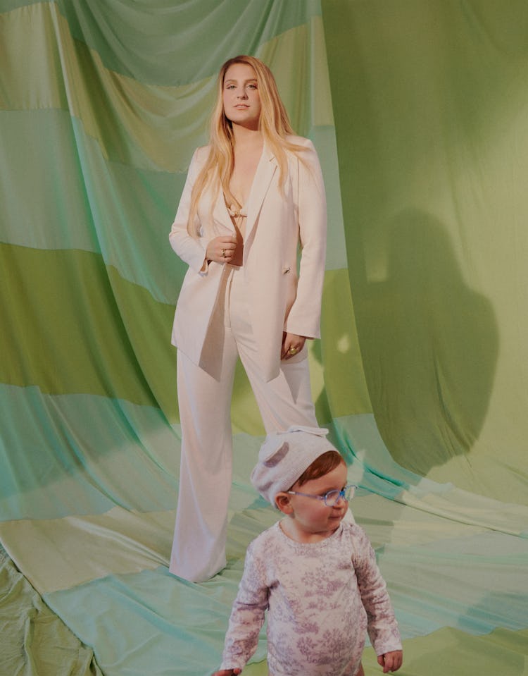 Meghan Trainor poses in front of green curtains, her son is in front of her looking to the side, bot...