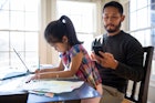 一位父亲盯着他的手机，而他的女儿在桌子旁做作业。