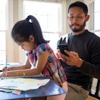 一位父亲盯着他的手机，而他的女儿在桌子旁做作业。
