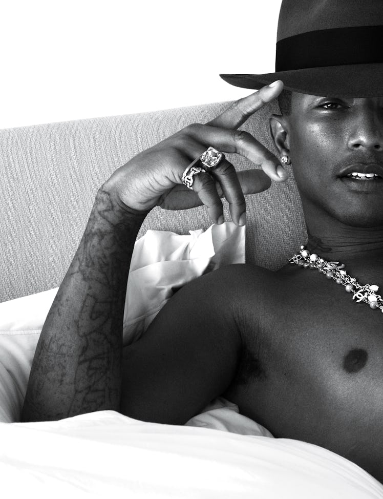Pharrell Williams lying in bed with a hat and chains on his neck in black and white