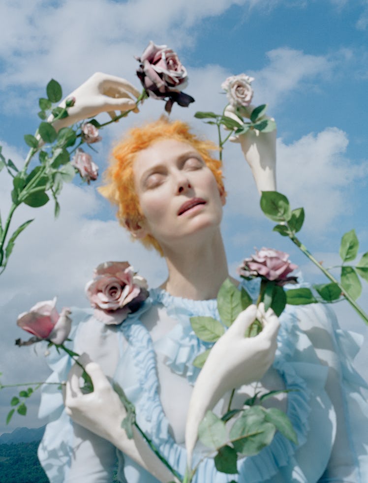 Tilda Swinton in a blue dress with flowers and white hand sculptures forming a circle around her fac...