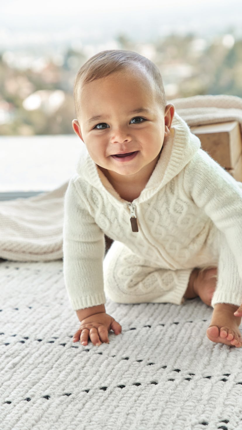 A baby on their hands and knees smiling at the camera, wearing a sweater onesie from the new Hilary ...
