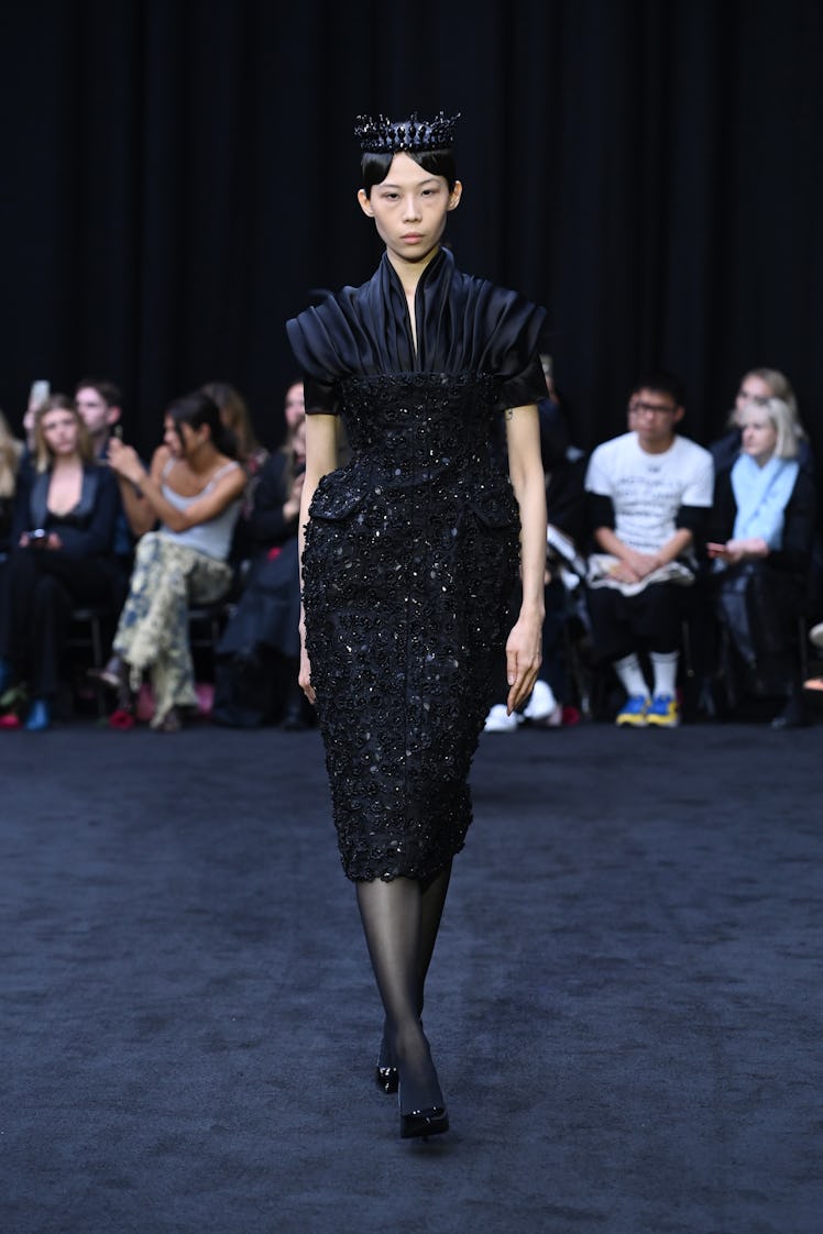 A female model walking the Richard Quinn spring 2023 runway while wearing a black dress and black he...