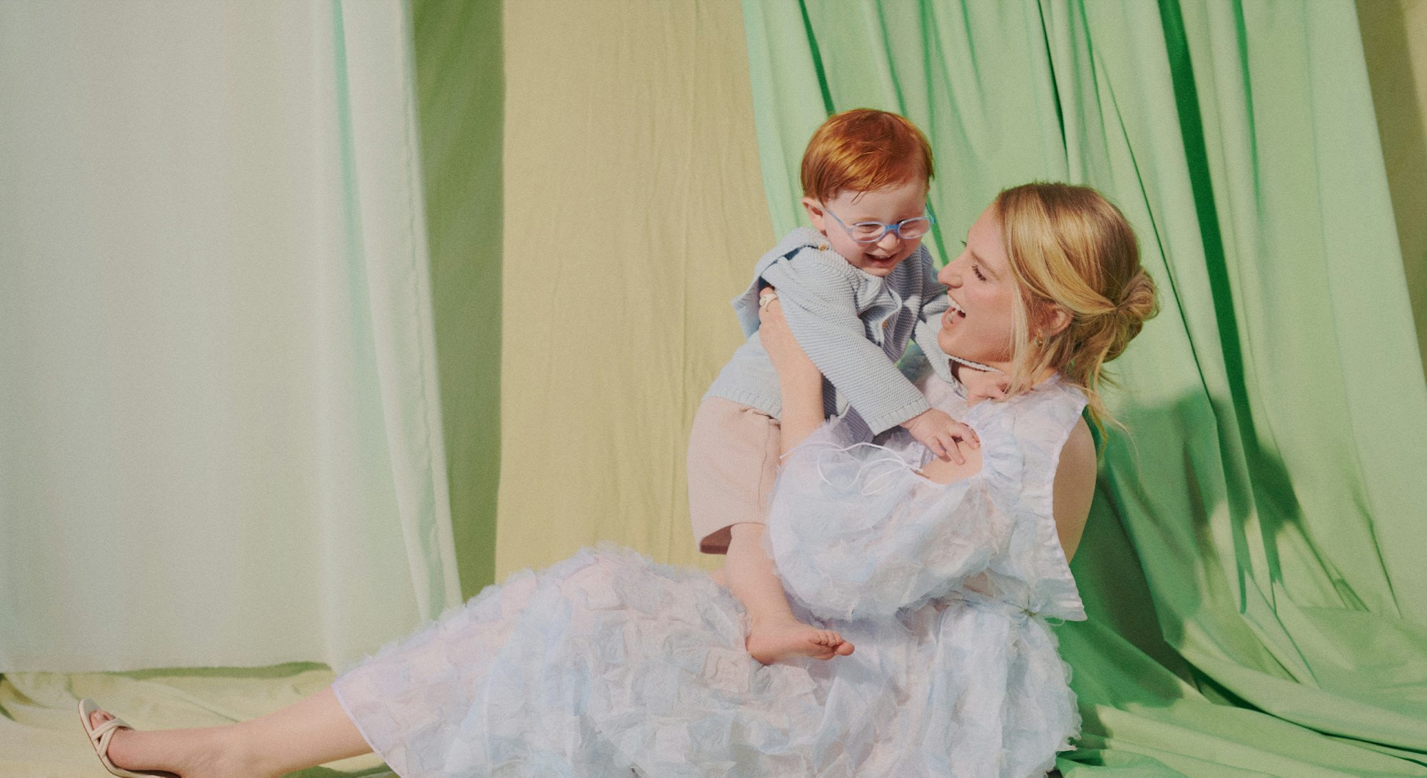 Meghan Trainor sitting on the ground in a white dress, playfully holding her son with green curtains...