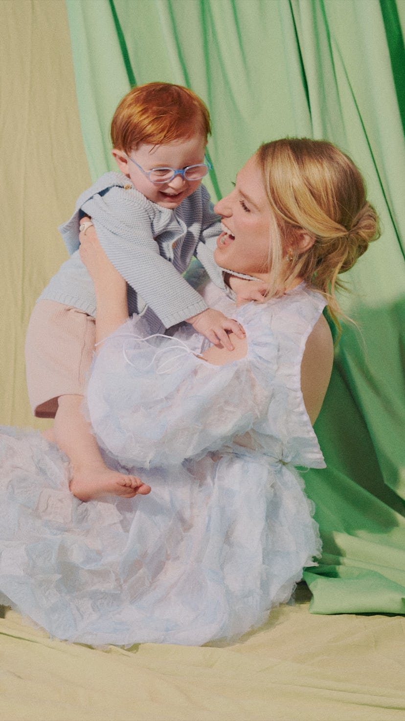 Meghan Trainor sitting on the ground in a white dress, playfully holding her son with green curtains...