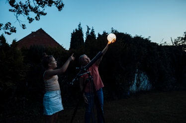 Older woman holding a lantern next to young girl in front of telescope, both looking to the sky..