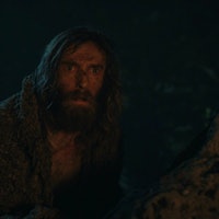 The Stranger (Daniel Weyman) crouches next to a rock in The Lord of the Rings: The Rings of Power Ep...