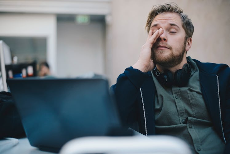 A sleep-deprived man rubs his eyes at work while on his laptop.