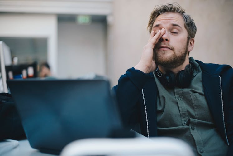 A sleep-deprived man rubs his eyes at work while on his laptop.