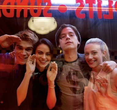 Cast of Riverdale standing behind glass looking straight ahead and smiling.