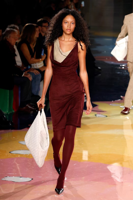 A model walks the Bottega Veneta Spring 2023 runway in burgundy dress, leggings and white maxi bag.