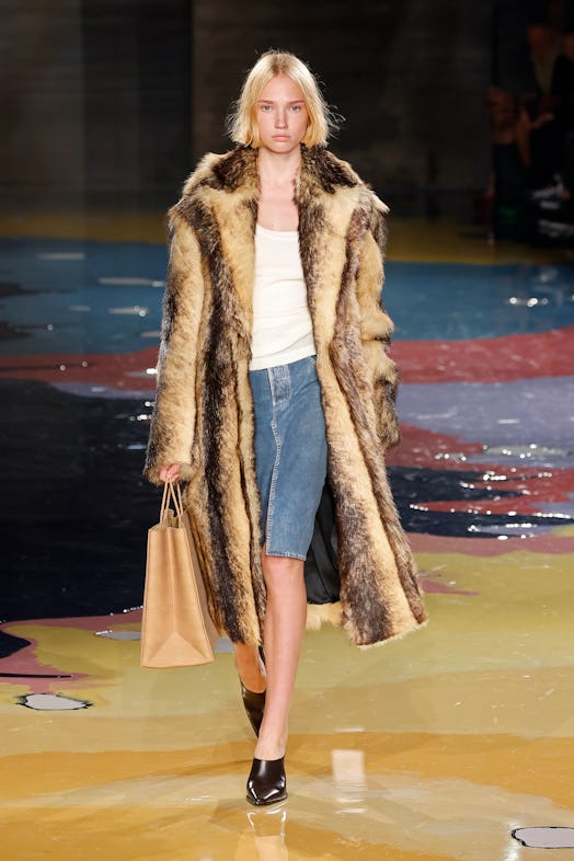 A model walks the Bottega Veneta Spring 2023 runway in a fur coat, white t-shirt, and denim skirt.