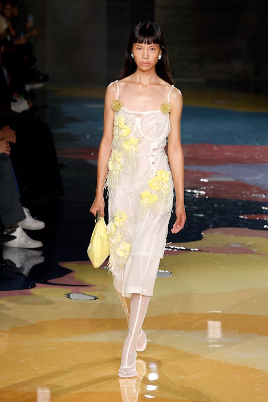 A model walks the Bottega Veneta Spring 2023 runway in a white dress with yellow flower details.