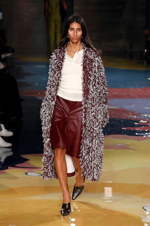 A model walks the Bottega Veneta Spring 2023 runway in leather burgundy skirt, white shirt and furry...