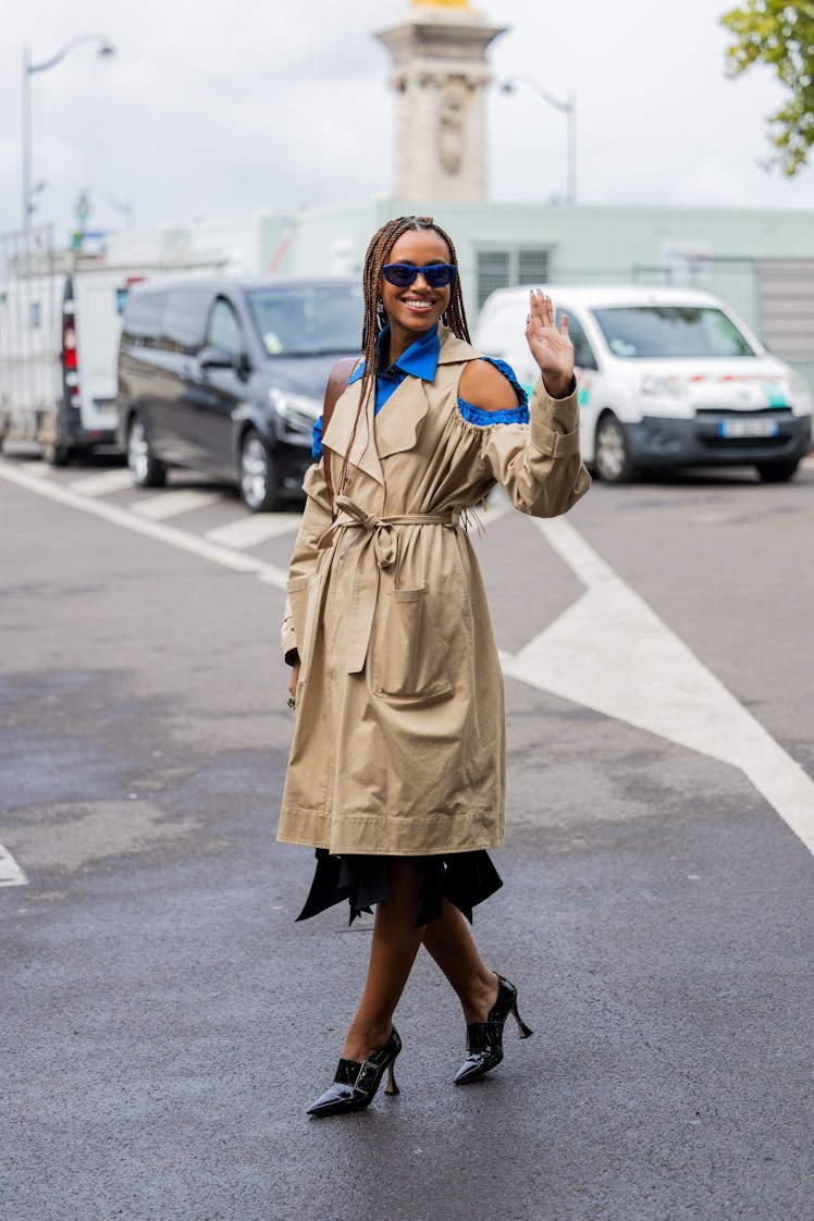 A guest wears cut out beige trench coat, cut cut blue blouse, black skirt, heels outside Victoria/To...