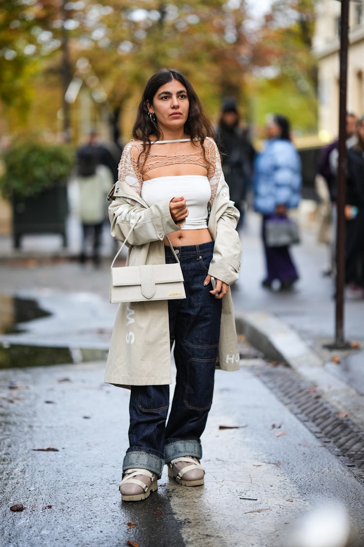 A guest wears silver earrings, a beige long coat, a white shoulder-off / cropped top, a brown fishne...