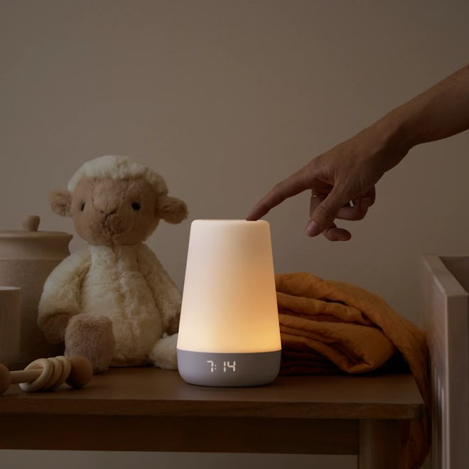 The Hatch sound machine and night light rests, illuminated, on a bedside table with a stuffed lamb.