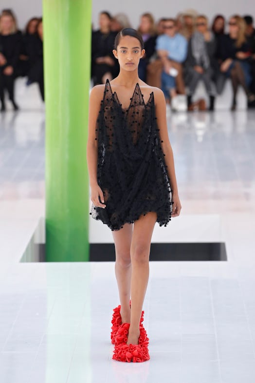 A model walking the runway in a laced short black dress and red flower shoes at Loewe Spring 2023 Pa...