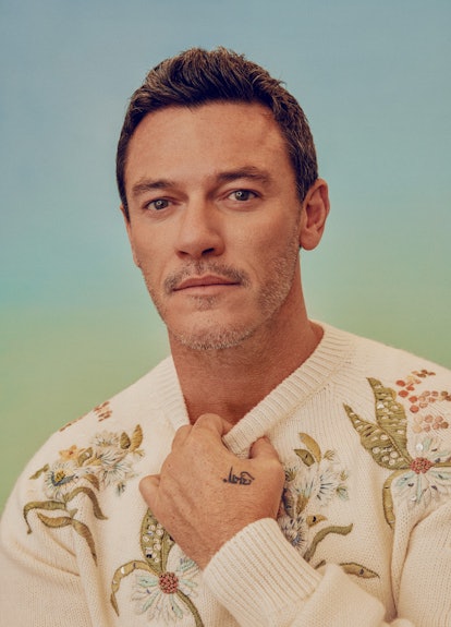 Luke Evans posing in a white Dior Men sweater with flowers on it, slightly pulling on the collar and...