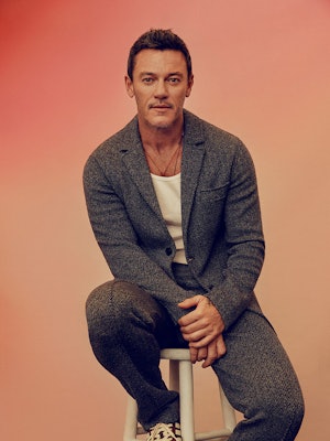 Luke Evans sitting on a high chair in a Missoni jacket and pants, a Calvin Klein tank top and Adidas...