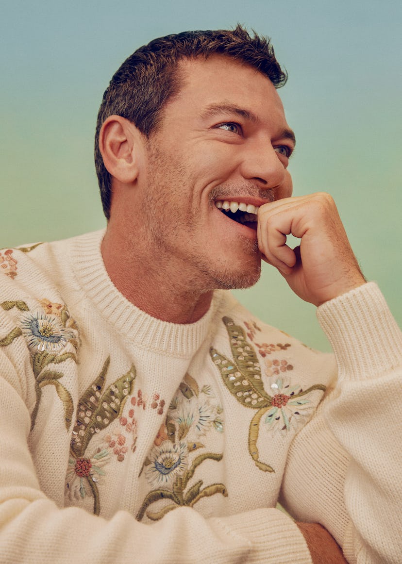 Luke Evans laughing in a white, flower-motived, Dior Men sweater