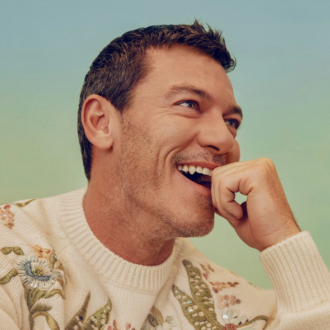 Luke Evans laughing in a white, flower-motived, Dior Men sweater