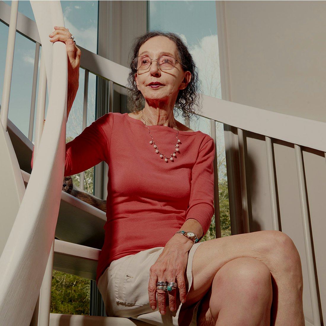 Joyce Carol Oates sitting on stairs in a coral shirt and beige skirt 
