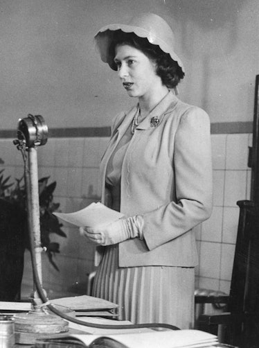 Queen Elizabeth wearing a statement hat and standing at a microphone