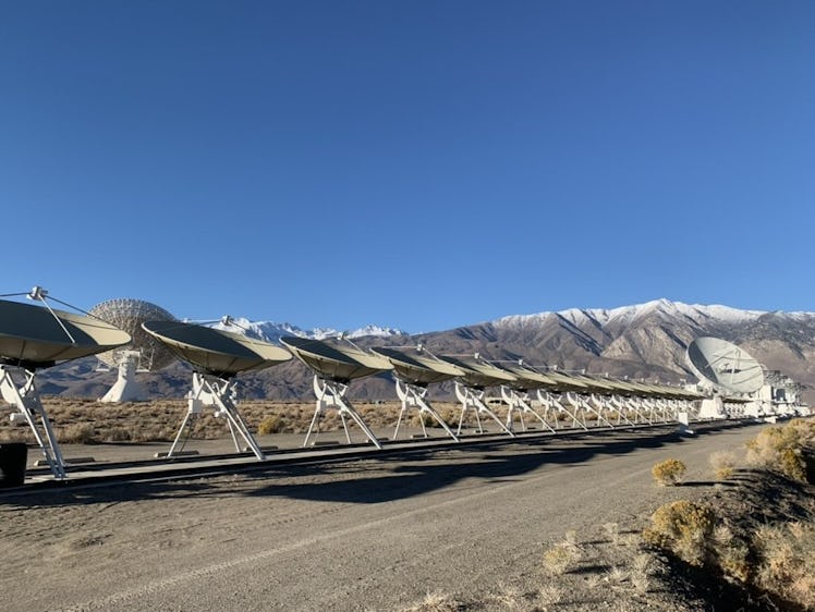 COlor photo of a long line of radio dishes on the desert floor with mountains in the background and ...