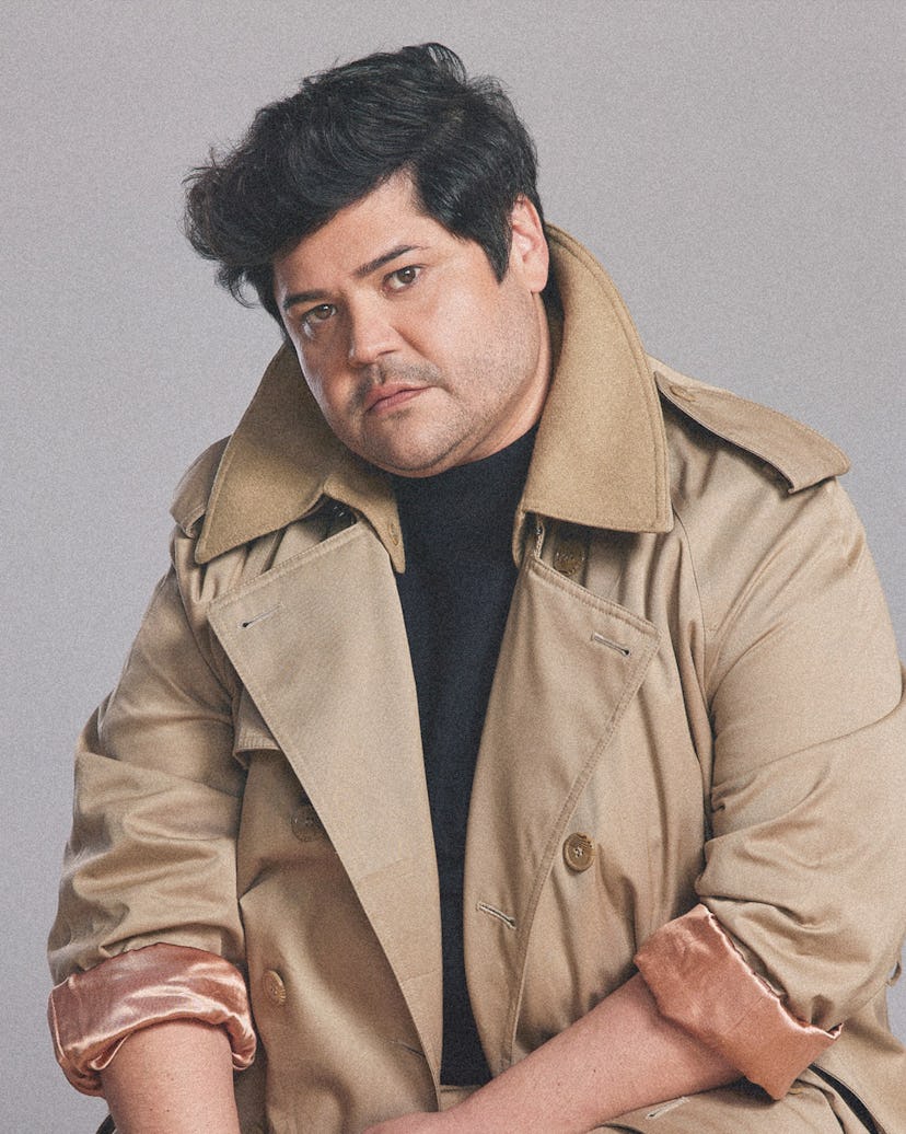 A portrait of Harvey Guillén in a black shirt and beige trench coat